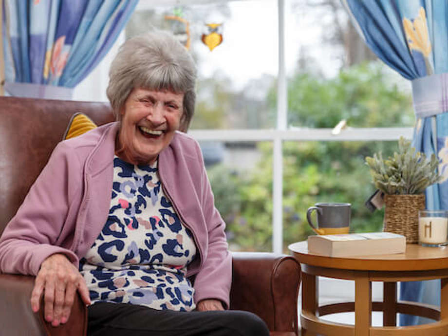 A woman sitting on an armchair in a communal lounge is belly laughing