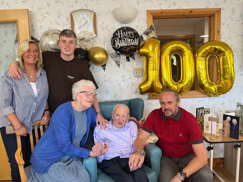 Ronnie sits in the centre of the room with his family surrounding him. 100th birthday celebration balloons, bunting, and cards are present throughout the scene