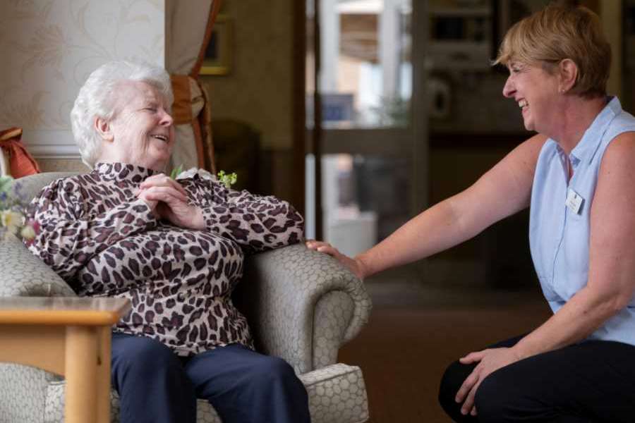 A staff member kneeling down to converse with a woman in a beige chair, they are both smiling and facing one another