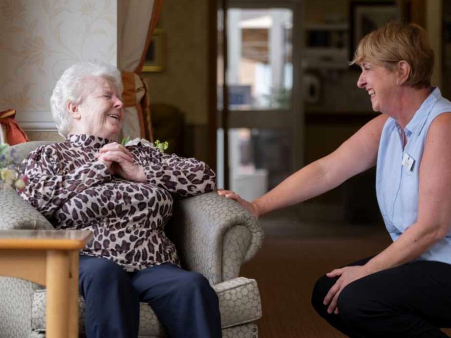 A staff member kneeling down to converse with a woman in a beige chair, they are both smiling and facing one another