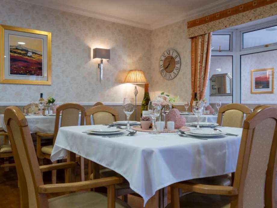 A dining room set up with multiple tables and chairs, napkins, plates, cutlery and fresh flowers throughout. There is art on the walls, with windows to the rear