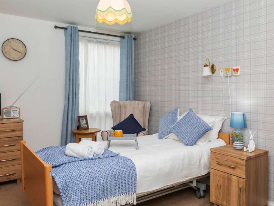 This comfortable bedroom set up compromises of a bedside table, bed, chair, and chest of drawers. The decor is neutral, with pops of blue throughout. There are fluffy towels on the bed and a range of personal belongings giving it a cosy feel