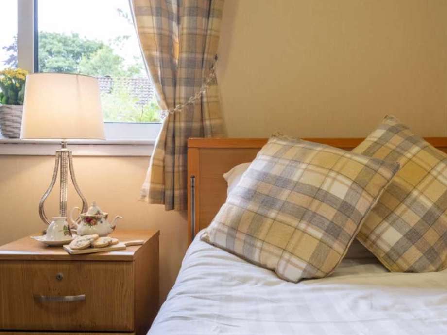 Decorative bedroom set up with a bed and side table. A window and the curtains and just in view, with a lamp and china tea set below