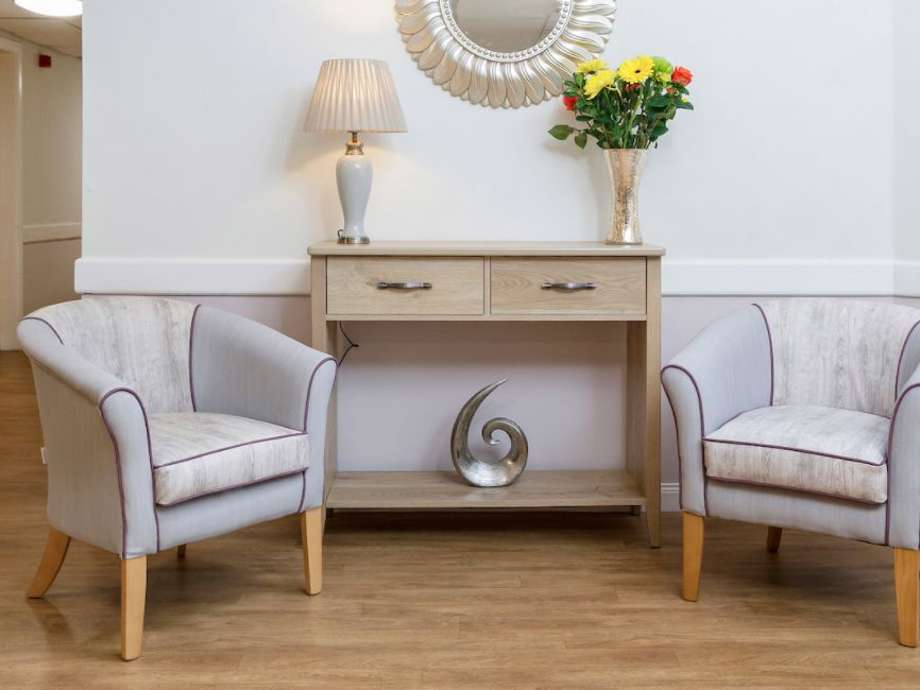 care home entrance: there are two grey seats with a wooden sideboard inbetween adorned with a lamp and fresh flowers