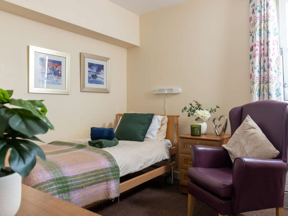 Milford House bedroom: An armchair and a bed within a cosy, spacious room full of artwork, flowers, and homely cushions. 