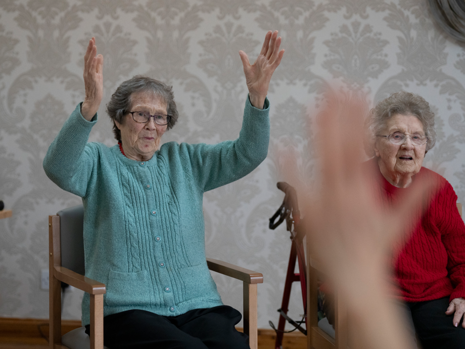 one woman in a blue jumper and one in a red knit copying a yoga instructors movements, their hands are in the air