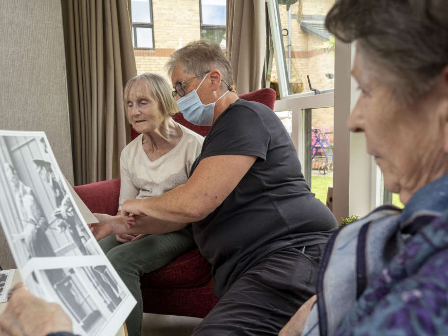 two care home residents and a staff member are sitting in the communal lounge area next to the window. They are holding old photographs, reminiscing on historic events