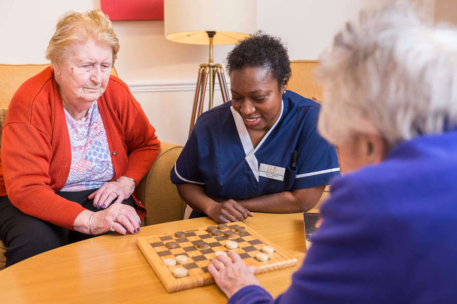 three people enjoy a game of checkers. Two people sit on armchairs, while the other crouches down to the table 