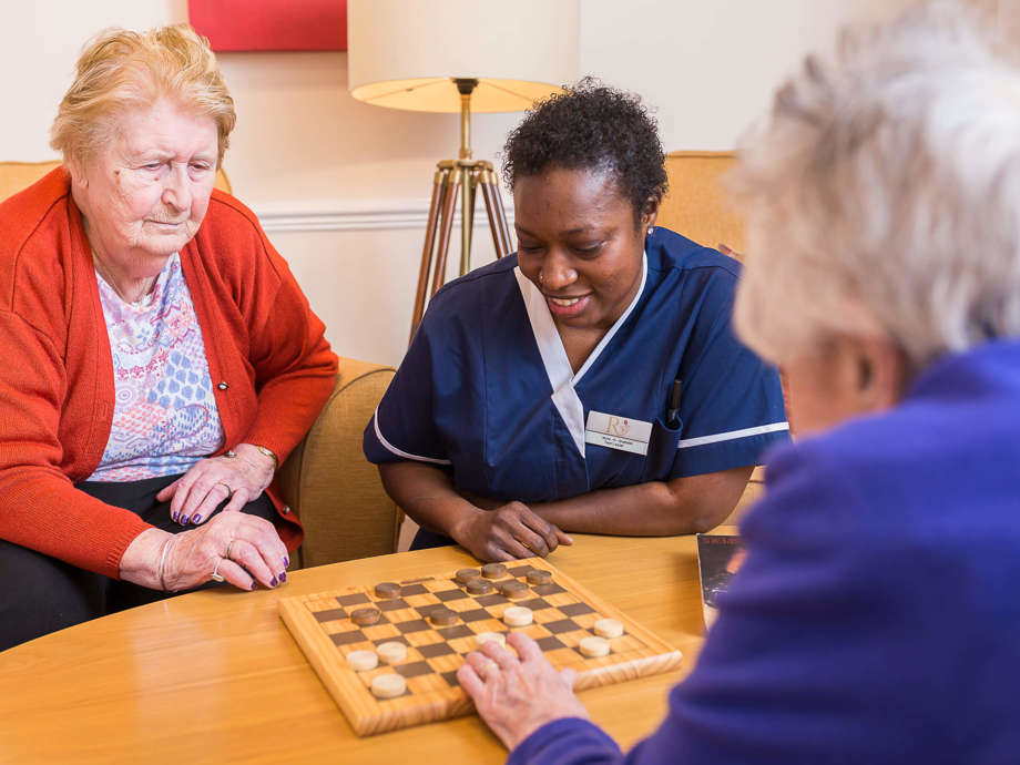 three people enjoy a game of checkers. Two people sit on armchairs, while the other crouches down to the table 