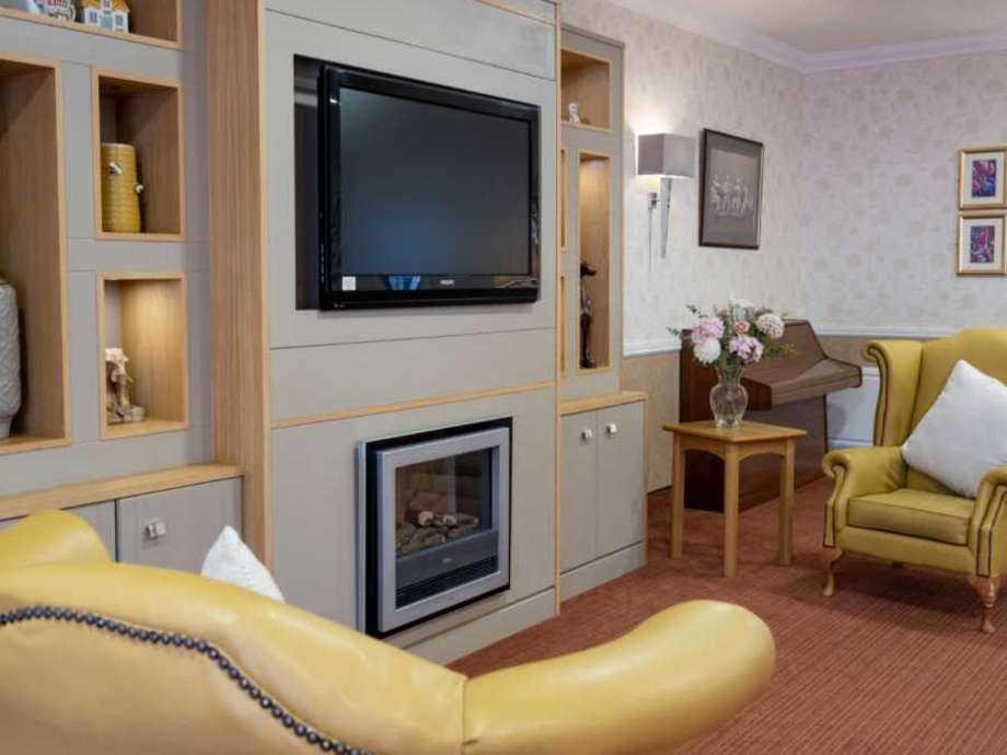 A communal lounge area with a TV set within a media wall at the centre. There is a fireplace set below the tv, with art on the walls and a piano in the background. Two yellow armchairs are in sight