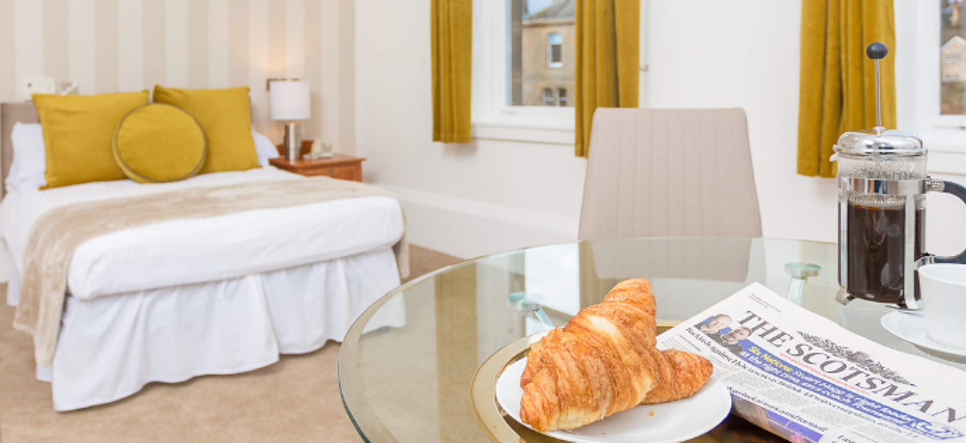 Airy bedroom with large windows offers ample light and space for relaxation. Includes dining table, bed with linens, and breakfast spread.