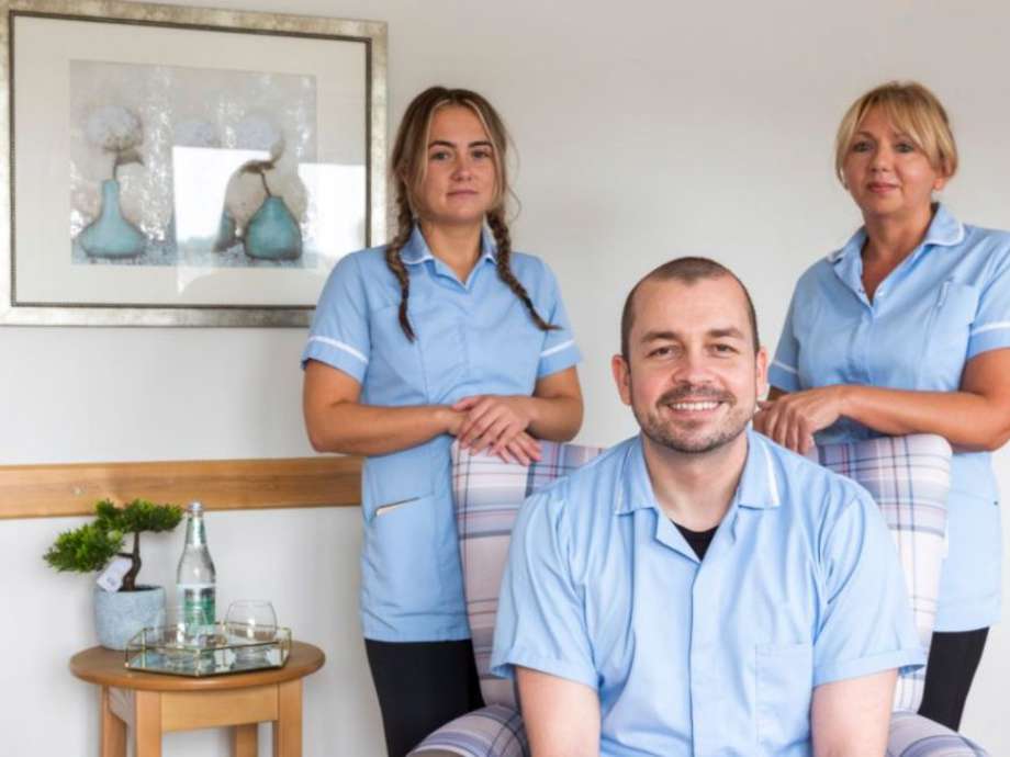 Two staff members stand behind a third staff member who is sitting in an armchair within the homes communal lounge. The environment is modern and bright