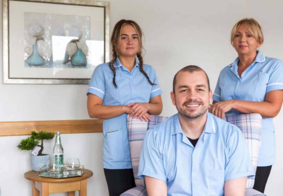 Two staff members stand behind a third staff member who is sitting in an armchair within the homes communal lounge. The environment is modern and bright