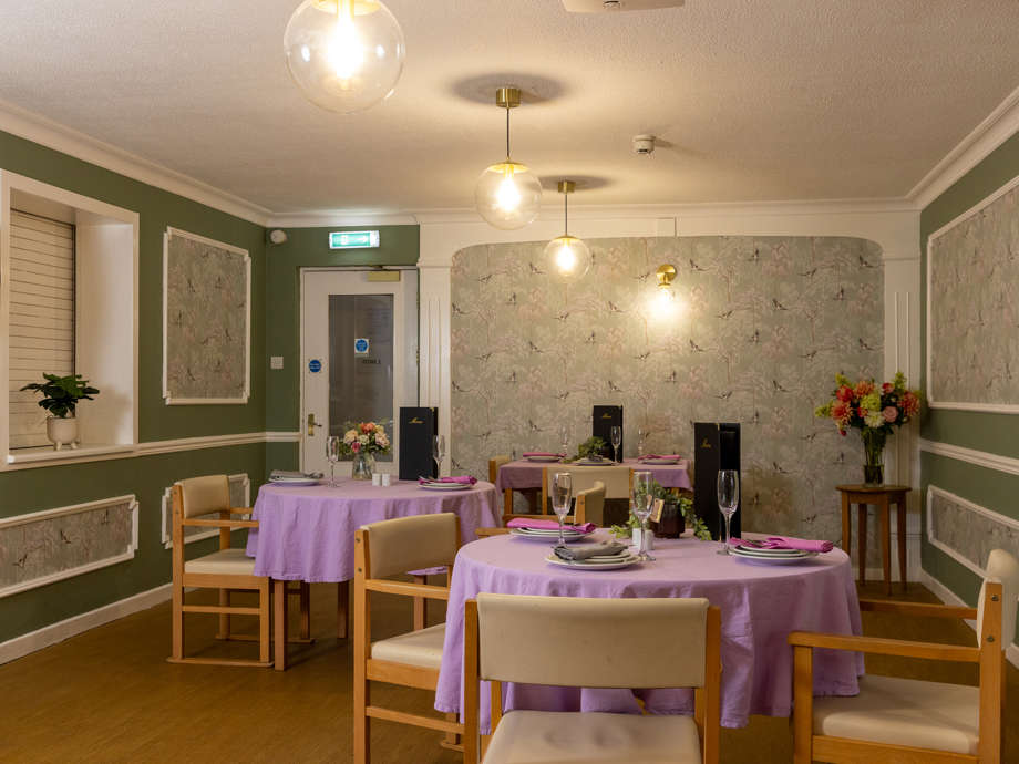 Dining tables and chairs set up for dinner service. There are purple tablecloths, glassware and pendant lighting 