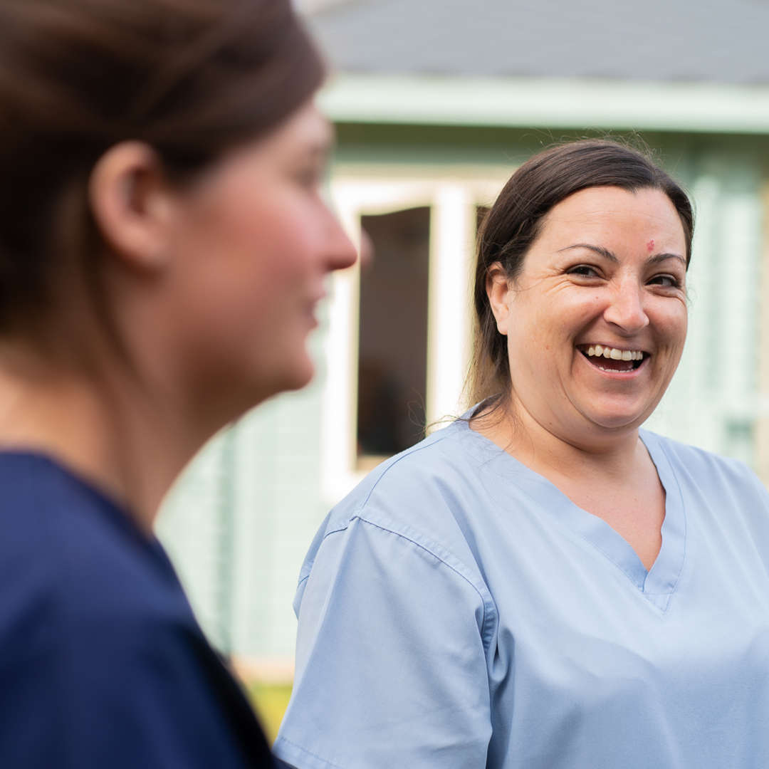 Two staff members stand in the garden. Only one is in focus, she is looking over and laughing