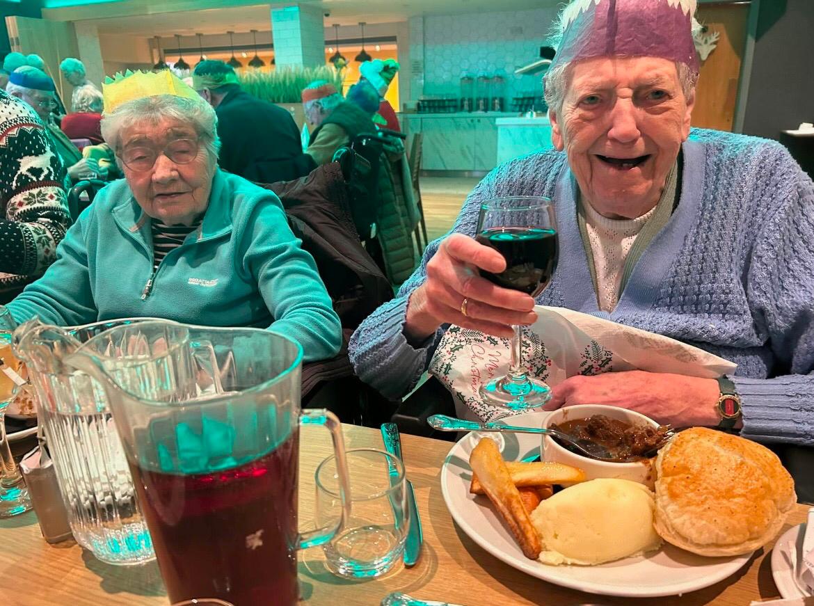 Care Home residents enjoy a Christmas meal together, one lifts her glass of wine up in a 'cheers!' motion