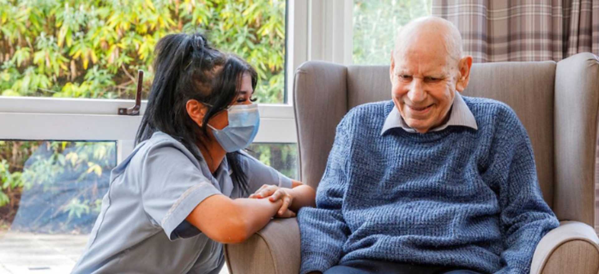 A staff member kneeling down to converse with a gentleman in a grey chair next to a large window looking out to the garden