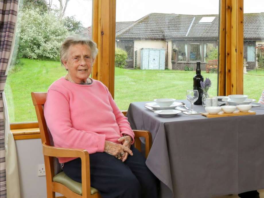 A care home resident sits in a wooden chair at a table in a care home dining room. A large window behind her offers a view of the garden.