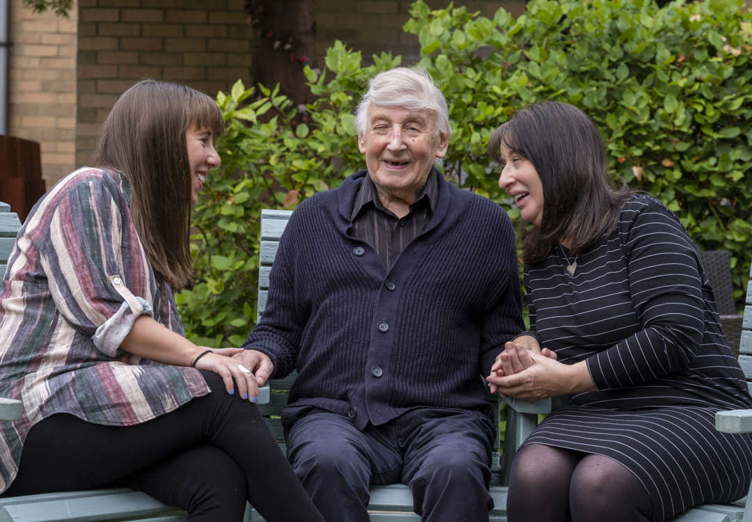 3 generations of a family are sitting in the garden on blue deck chairs as the family visit their father and grandfather respectively 