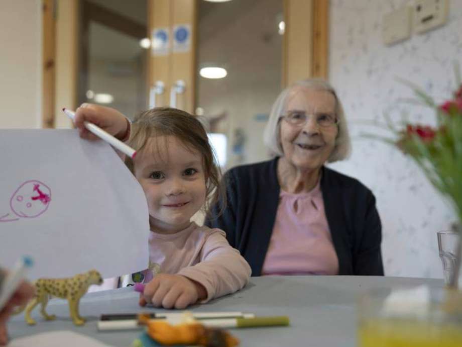 A nursery child holds up her latest drawing. A care home resident sits behind offering an encouraging look