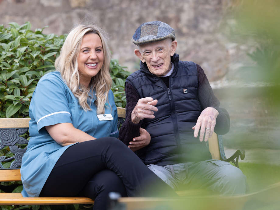 A staff member and a resident sit beside each other on a bench in the care homes garden. They are laughing together