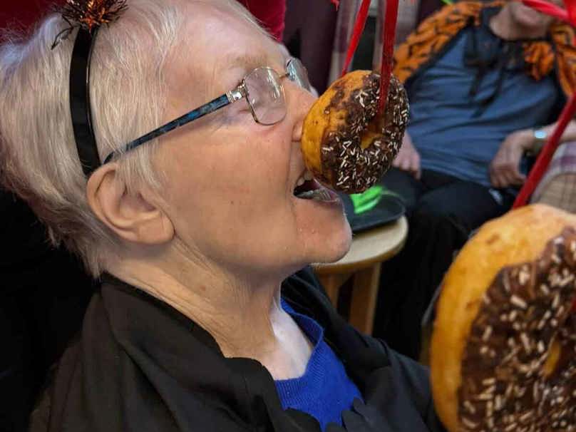 A care home resident reaches for a donut hanging from a string, dressed in a festive Halloween costume