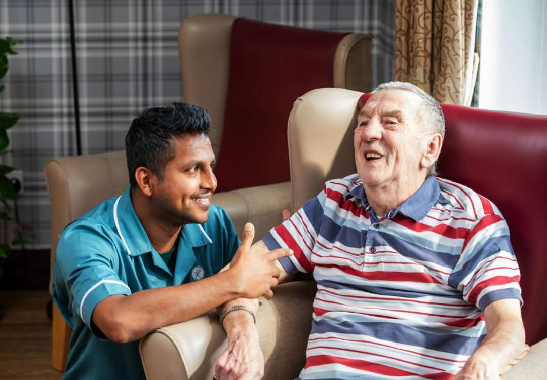 A staff member kneeling down to converse with a gentleman in a red/beige next to a large window looking out to the garden