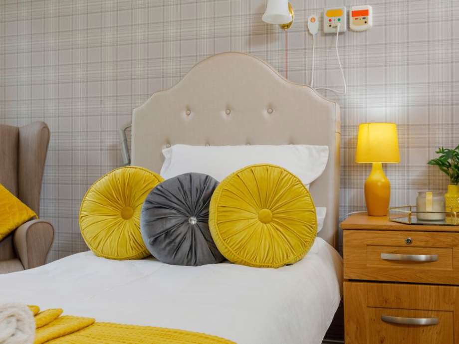 A bedroom set up from the home. Grey and beige tartan wallpaper lines the walls, with a neutral bed and chair and cosy yellow accents throughout