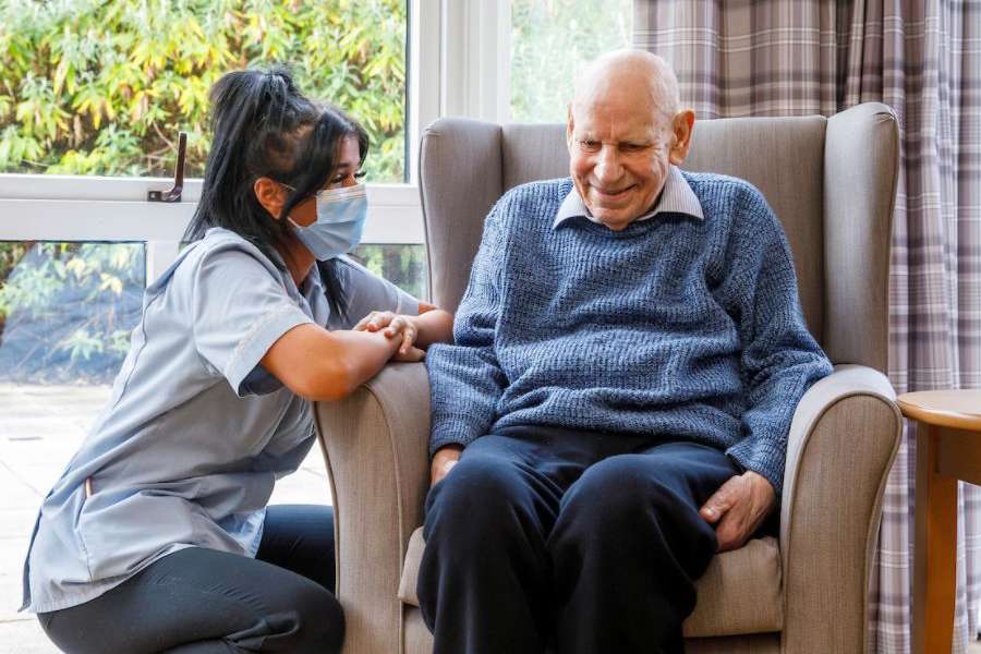 A staff member kneeling down to converse with a gentleman in a grey chair next to a large window looking out to the garden