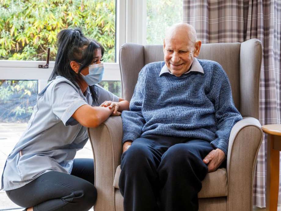 A staff member kneeling down to converse with a gentleman in a grey chair next to a large window looking out to the garden