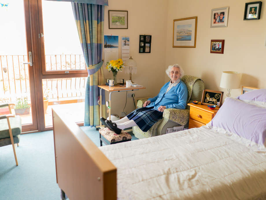 An older adult relaxes in a bedroom chair, enjoying a cup of tea and the view from the window