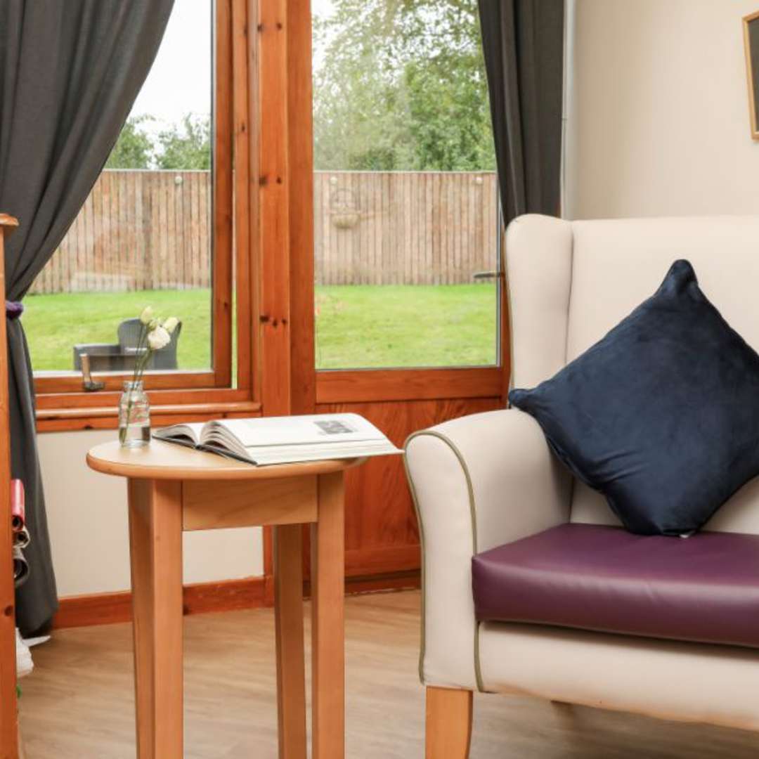 A cosy seating area in a care home. The nook has a chair with a navy blue pillow next to a small table holding an open book.