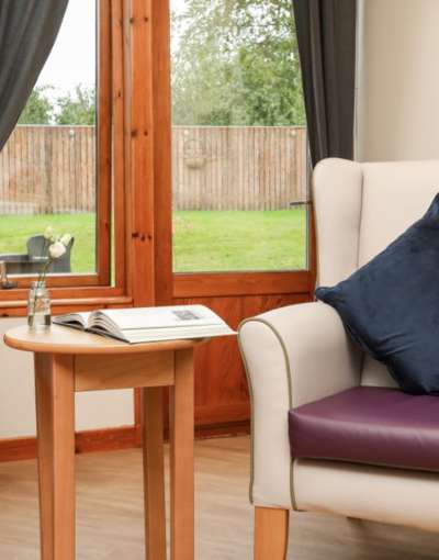 A cosy seating area in a care home. The nook has a chair with a navy blue pillow next to a small table holding an open book.