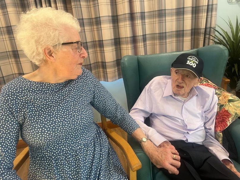 Married couple Eleanor & Ronnie sit side by side holding hands as they celebrate Ronnies 100th birthday