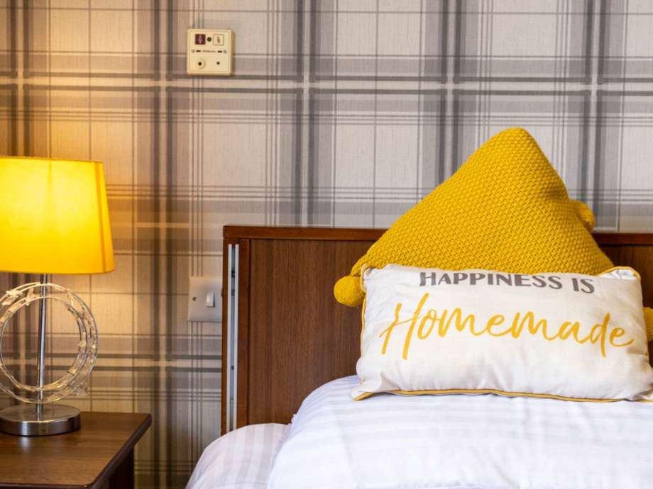 A closeup shot of a bedroom within the home, white duvet set with yellow cushions and a lamp on the side table