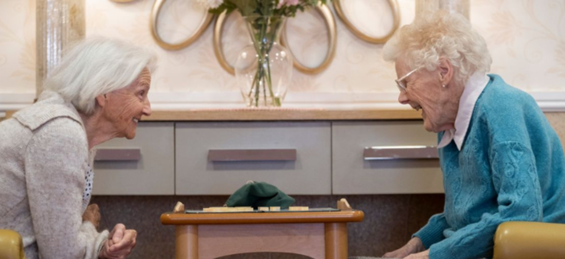 two residents sit across from one another, engaging in a game of scrabble. They are sitting on yellow armchairs, smiling at one another